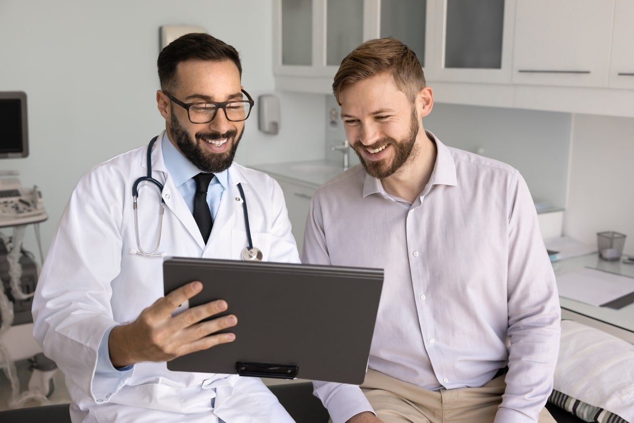 Happy male doctor and patient looking at good medical checkup results together, Positive Latin physician explaining examination report to patient, showing file on pad, smiling, laughing